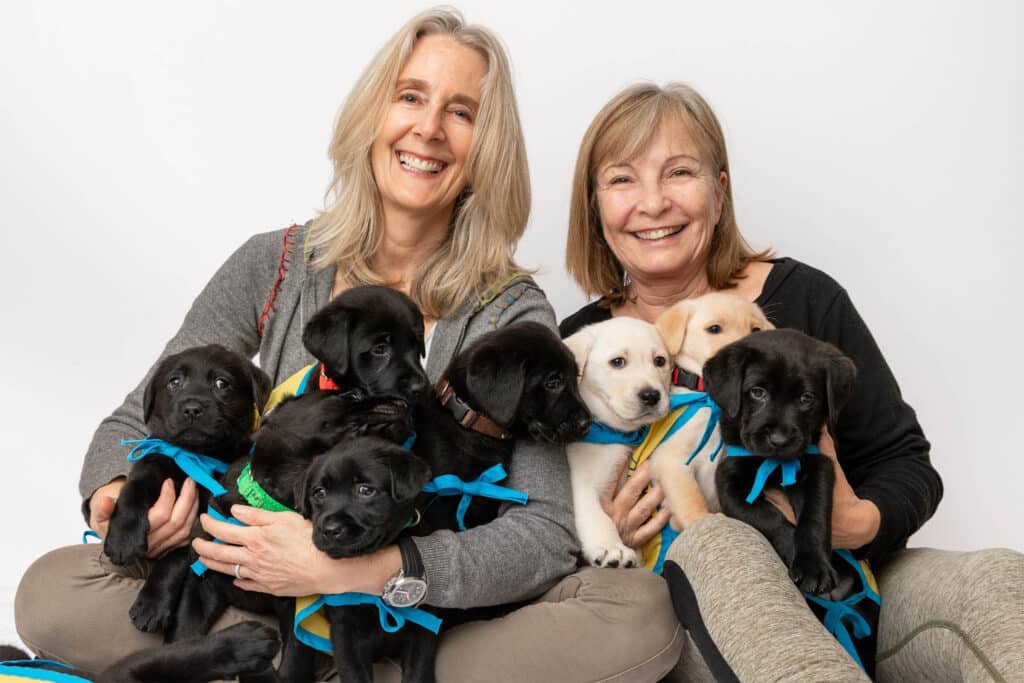 two smiling women holding a group of black and yellow lab puppies