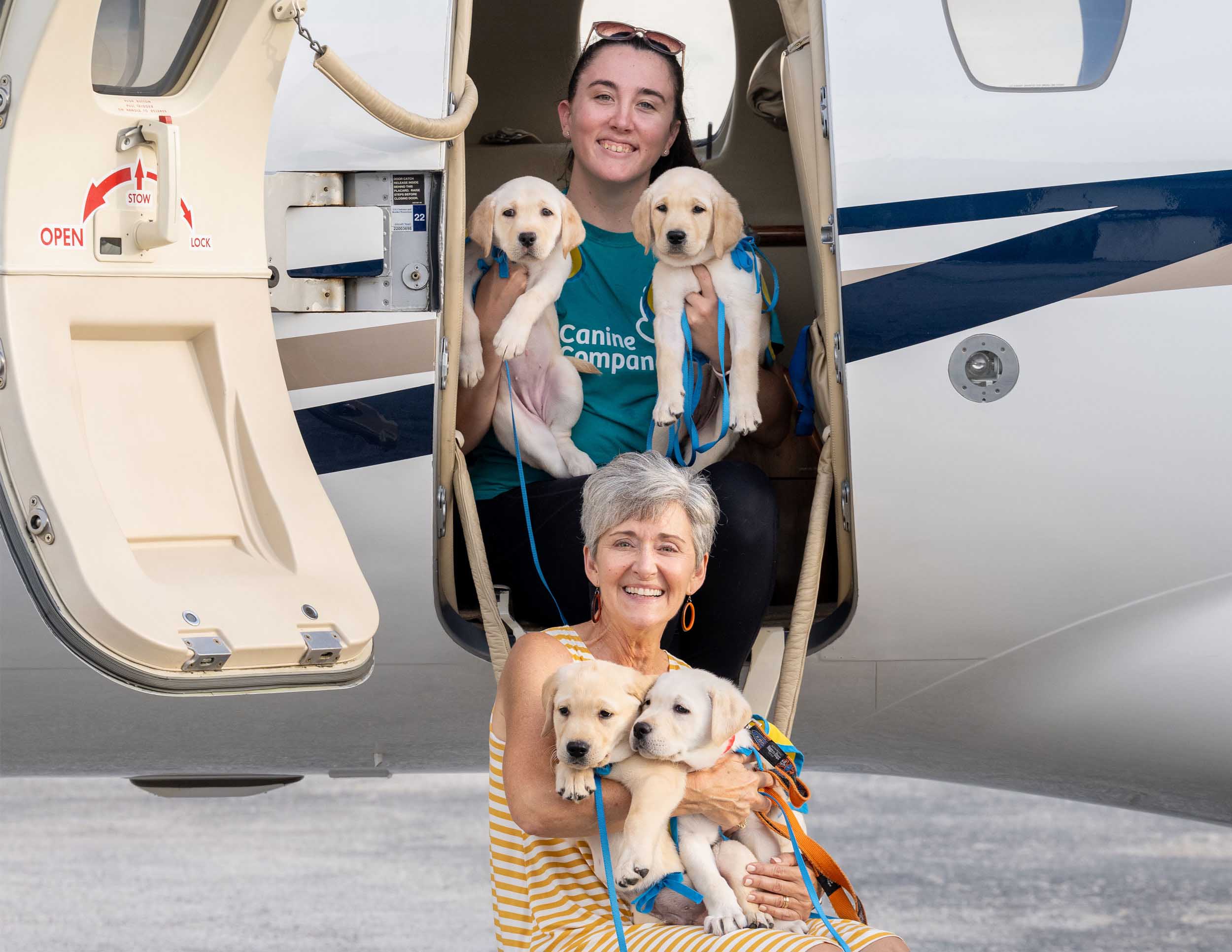two smiling volunteers holding puppies sitting in the doorway of a small plane