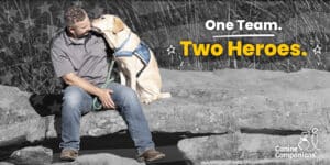 a smiling man sits on some rocks next to his sitting yellow lab service dog. The words below say One Team Two Heroes