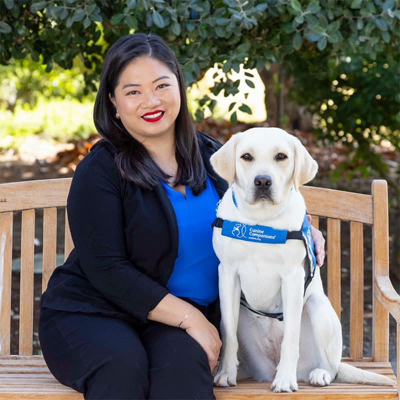 A smiling woman on a bench sitting next to a yellow lab service dog