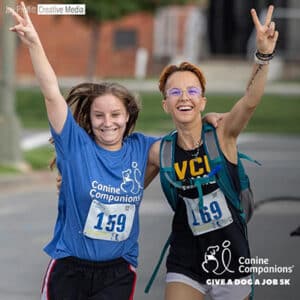 Two runners smiling and cheering during the Canine Companions "Give a Dog a Job" 5K race, wearing bib numbers 159 and 169.