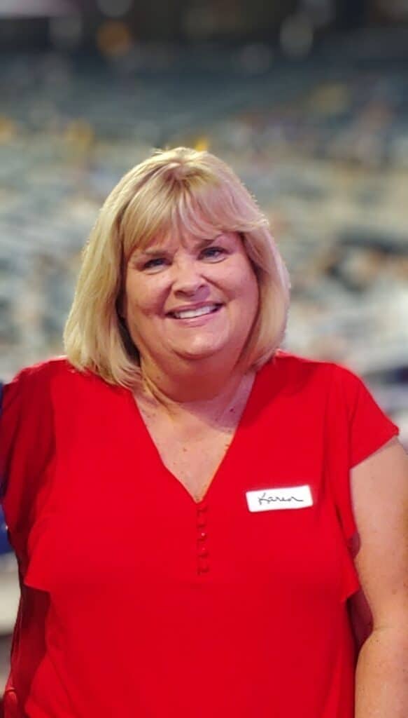 A smiling woman with blonde hair wearing a red top stands in front of a blurred background, possibly at an event or gathering. A name tag with "Karen" is visible on her shirt.