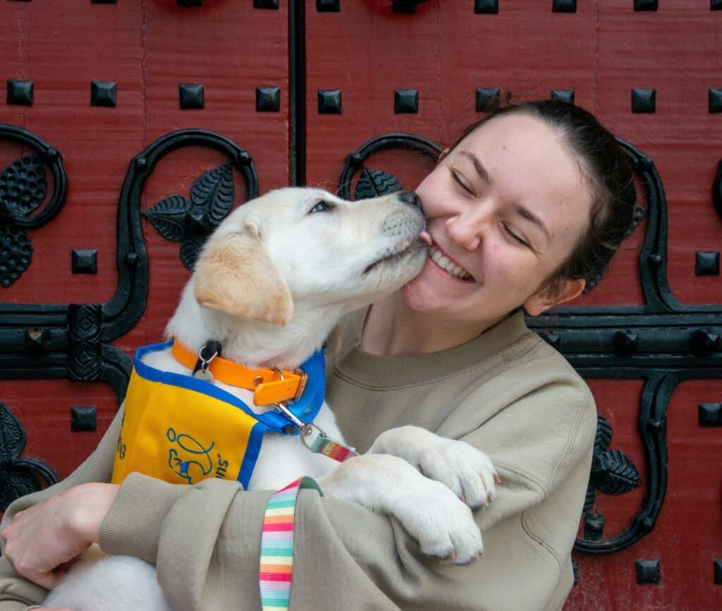 A woman smiles while holding a playful puppy that is licking her face, set against a decorative red door with black iron details.