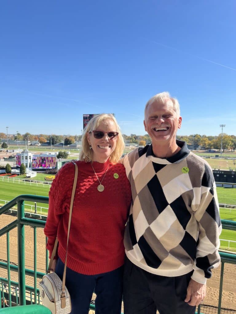 A woman in a red sweater and a man in an argyle-patterned sweater smile together at an outdoor racetrack under a clear blue sky. They are standing on a balcony with a grassy racetrack visible behind them.