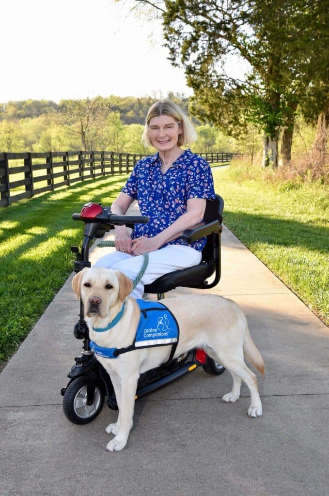 A smiling woman with short blonde hair sits on a mobility scooter beside a yellow Labrador dog wearing a harness that reads "Canine Companions." They are on a paved path with green grass and trees in the background.