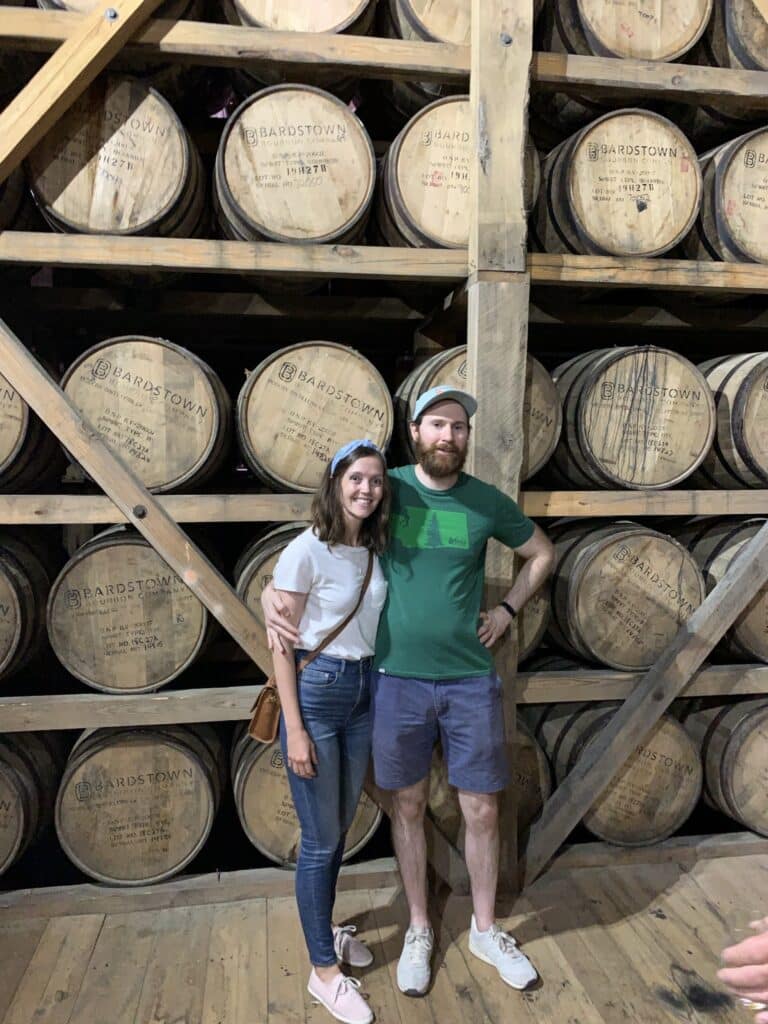 A man and a woman pose together in front of a wall of wooden barrels labeled "Bardstown," inside a rustic setting. The woman is wearing a white shirt and blue jeans, while the man is in a green shirt and shorts. They are smiling, with the woman's arm around the man's waist.