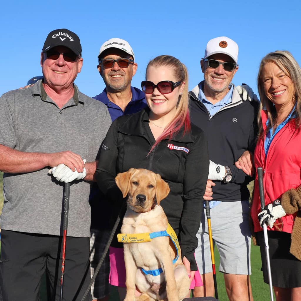 Three men, one woman and one young woman standing with a Labrador puppy wearing a yellow vest; they are all wearing outdoor attire, with two men and a woman holding golf clubs