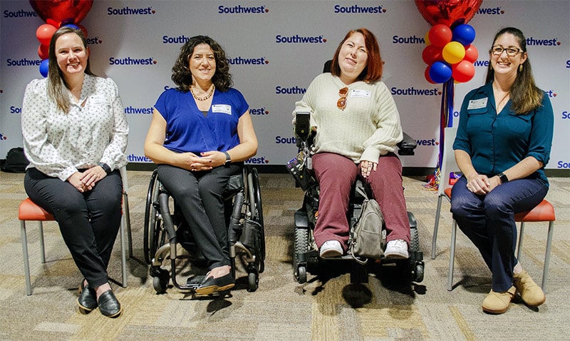 Four smiling women seated in a row at a Southwest Airlines event, two of whom are wheelchair users, in front of a backdrop with the airline's logo and decorated with balloons reflecting the company's colors.