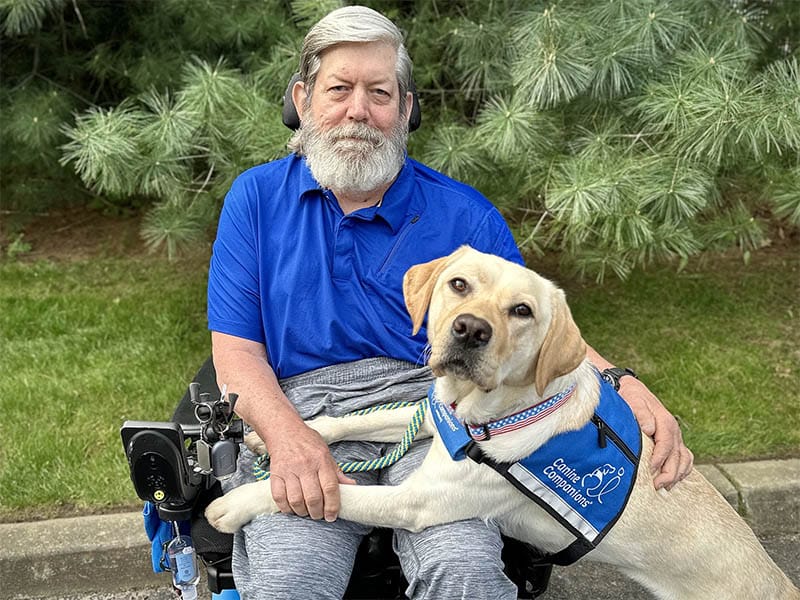 A man with a gray beard in a power wheelchair, joined by his devoted yellow Labrador service dog wearing a blue Canine Companions vest, poses together in front of a backdrop of lush pine trees.