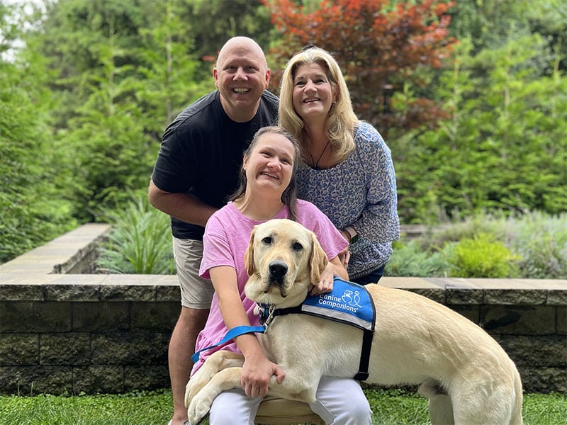A family of three, with a man, a woman, and a seated young woman, shares a joyful moment with a gentle yellow Labrador service dog in a lush garden setting.
