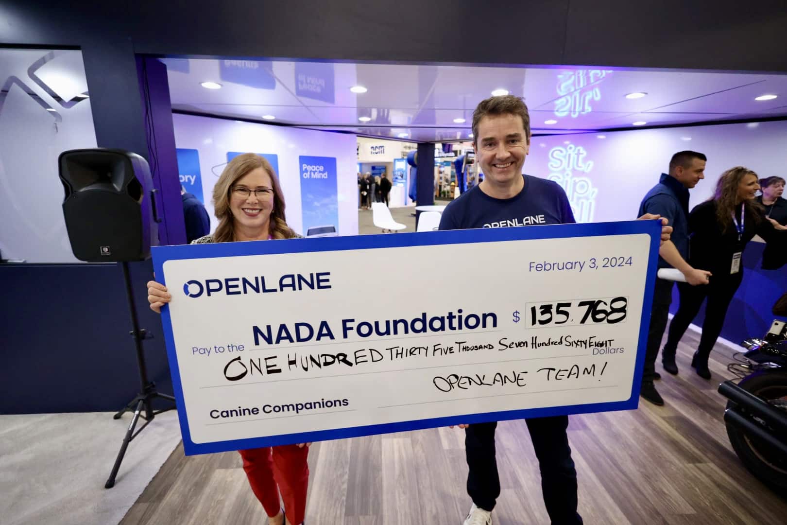 Two smiling individuals proudly display a large ceremonial check from OPENLANE to the NADA Foundation for $135,768, benefiting Canine Companions, dated February 3, 2024.