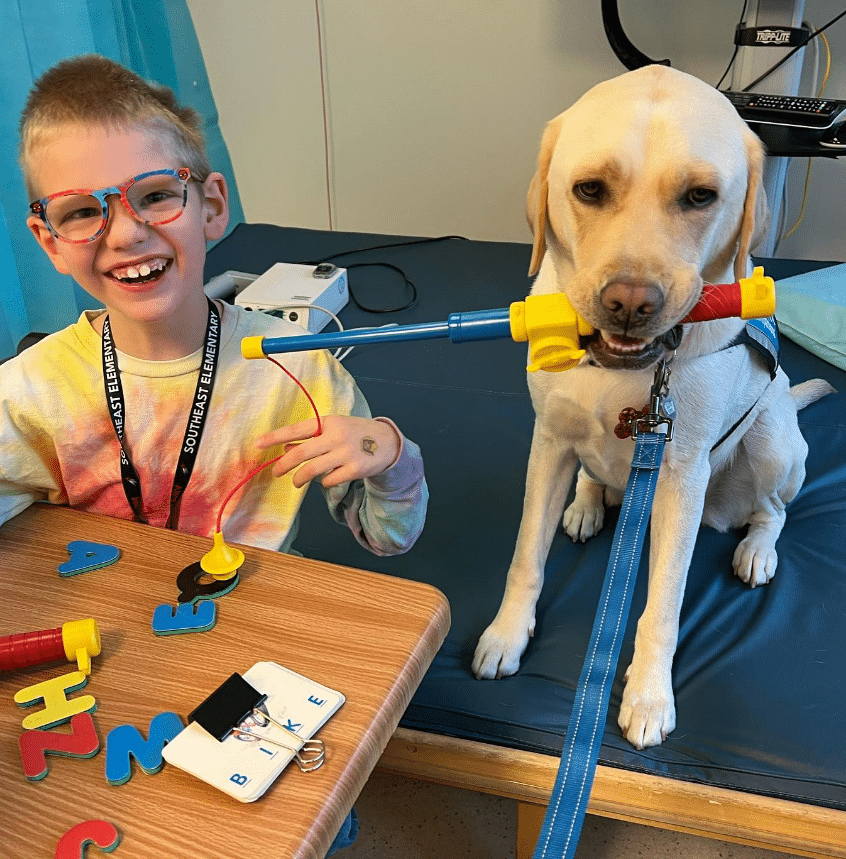 A boy with bright glasses smiles widely while a yellow Labrador service dog, holding a helping tool in its mouth, assists him at a table with educational toys.