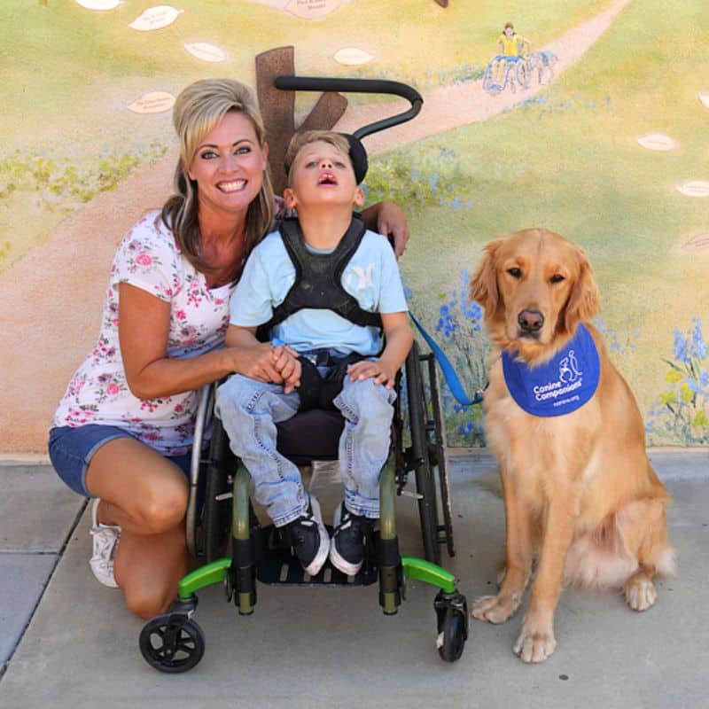 a smiling mother next to her son in a wheelchair with their golden retriever service dog in a blue canine companions bandana