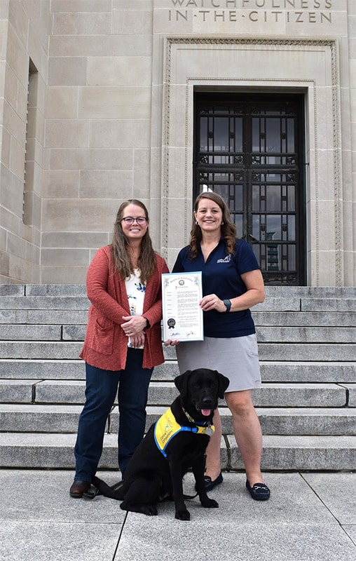 Two women stand on the steps of a stately building, smiling as they hold a certificate. A black Labrador service dog in a blue and yellow vest sits proudly in front, completing this moment of achievement.