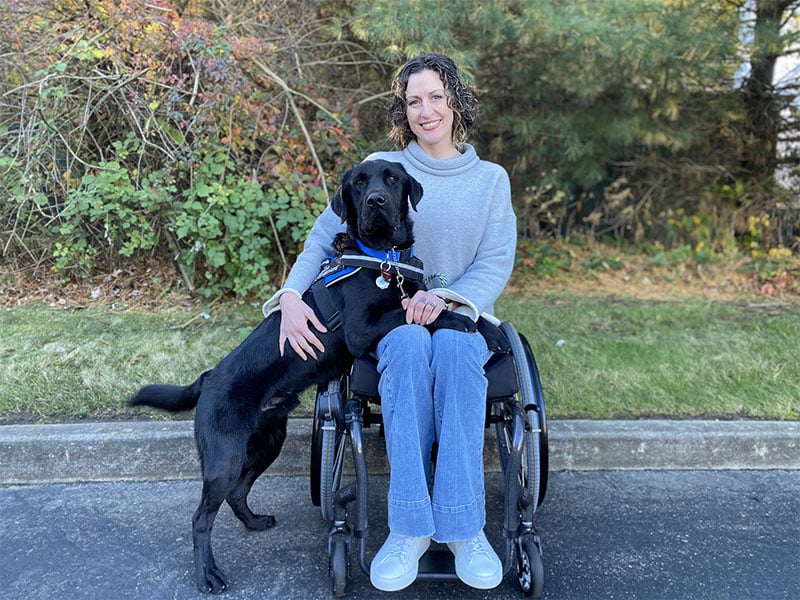 A smiling woman in a wheelchair accompanied by her attentive black Labrador service dog, both posing for the photo with a backdrop of trees and shrubs.