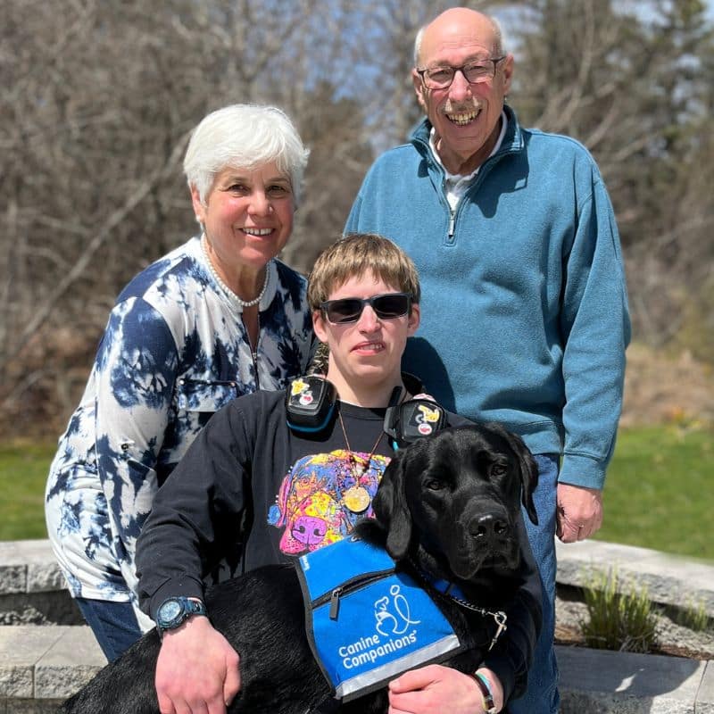 A family portrait in an outdoor setting: a young man wearing sunglasses is joined by a black Labrador service dog in a blue vest, with an older couple standing behind them, all smiling and enjoying a sunny day.