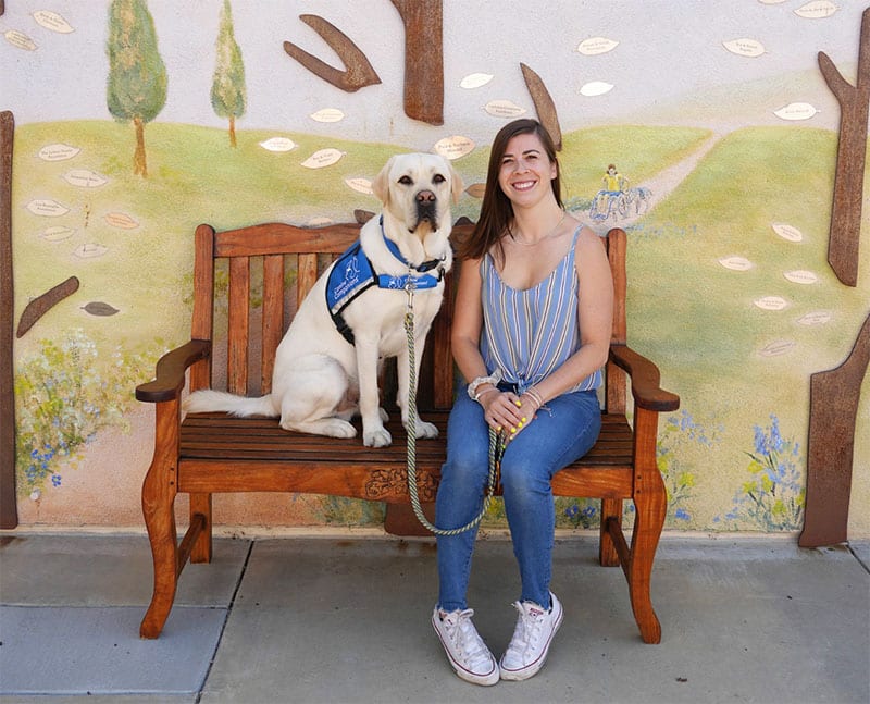 A cheerful woman sits beside her yellow Labrador service dog on a wooden bench. The dog, donning a Canine Companions vest, and its handler are in front of a whimsical mural depicting a pastoral landscape.