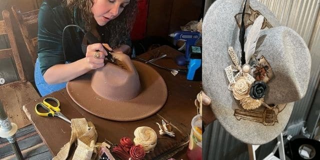 A focused craftswoman is meticulously shaping and detailing a brown felt hat with a hot glue gun, surrounded by various millinery tools and decorative items on a rustic wooden table next to A handcrafted gray felt hat adorned with a creative arrangement of decorative elements including lace, feathers, pearls, and a cluster of flowers, showcasing a vintage-inspired design.