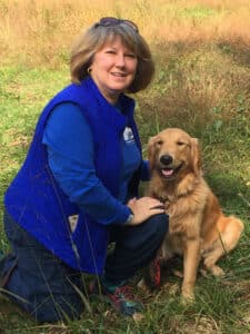 A woman in a blue vest kneels beside a smiling golden retriever in a field, both looking happy and content in the moment.