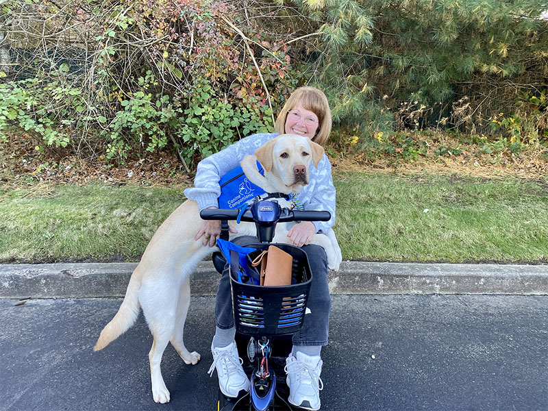 A smiling woman on a mobility scooter is affectionately joined by her loyal yellow Labrador service dog wearing a blue Canine Companions vest, outdoors with greenery in the background.