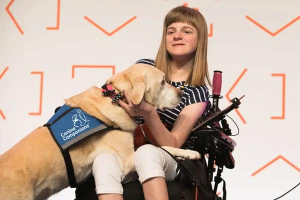 A young girl in a wheelchair shares a loving moment with her service dog, a golden Labrador wearing a Canine Companions vest, against a backdrop with orange patterns.