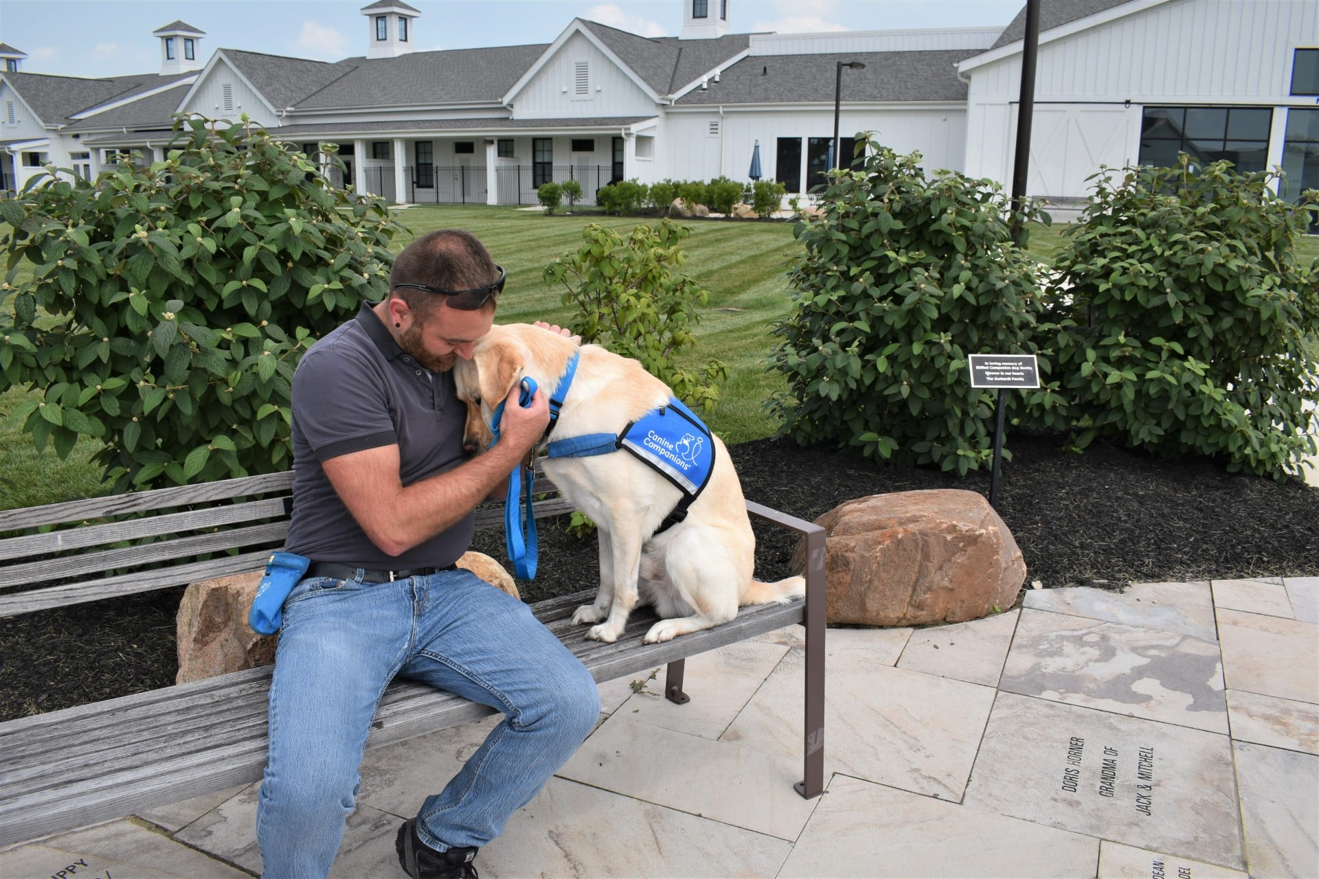 A man shares a tender moment with a yellow Labrador service dog on a bench, in front of a building with lush greenery around.