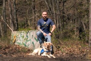 A man wearing a U.S. Marines tee shirt sits on a graffitied concrete wall in the woods. A yellow Labrador in a blue Canine Companions service dog vest lies at his feet looking at the camera.