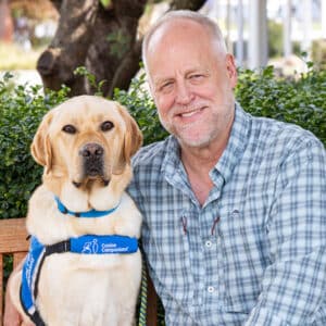 a smiling man sitting next to a yellow lab service dog
