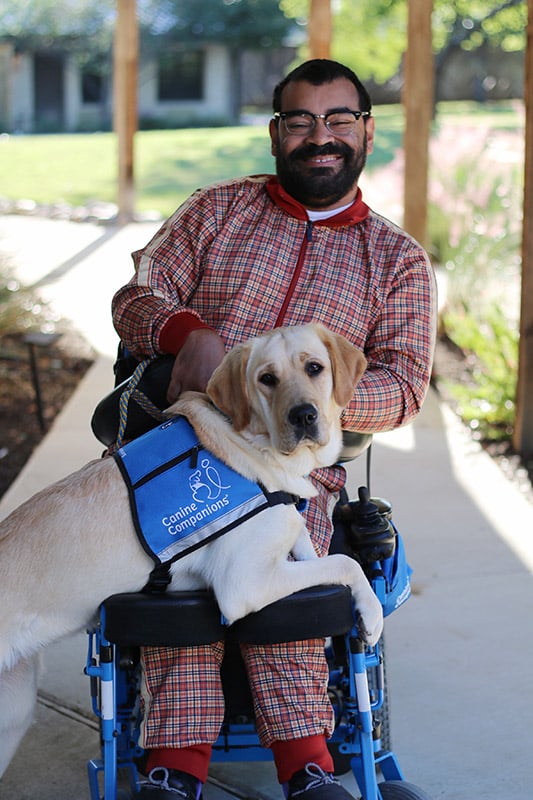 A man with a warm smile is seated in a wheelchair, accompanied by his loyal yellow Labrador service dog wearing a blue Canine Companions vest, both posing happily in an outdoor setting with a walkway and columns in the background.