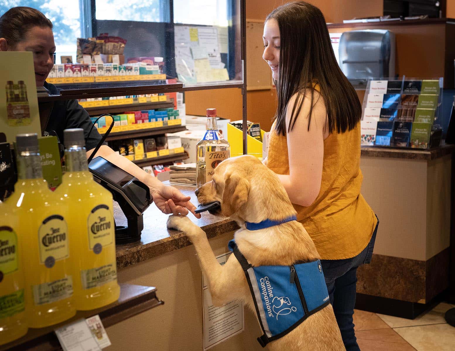 A woman at a cash register with her service dog, who is holding out a wallet to the cashier