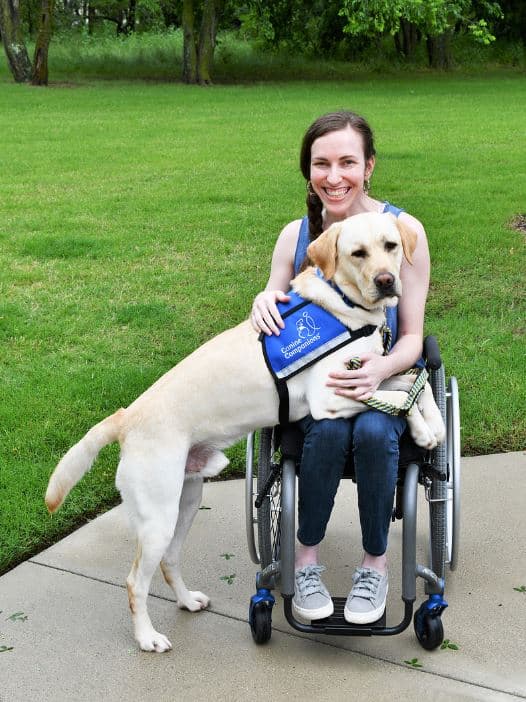 a smiling woman in a wheelchair with a yellow lab service dog standing with it's body across her lap