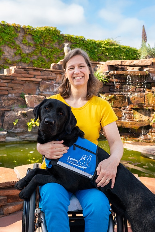 Woman in a wheelchair holding her black lab service dog by the fountain