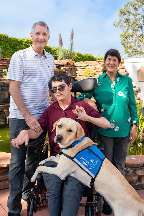 Young man seating in a wheelchair with his parent behind and his service dog