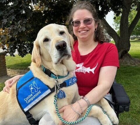 A smiling woman in a motorized wheelchair with a yellow lab service dog laying across her lap