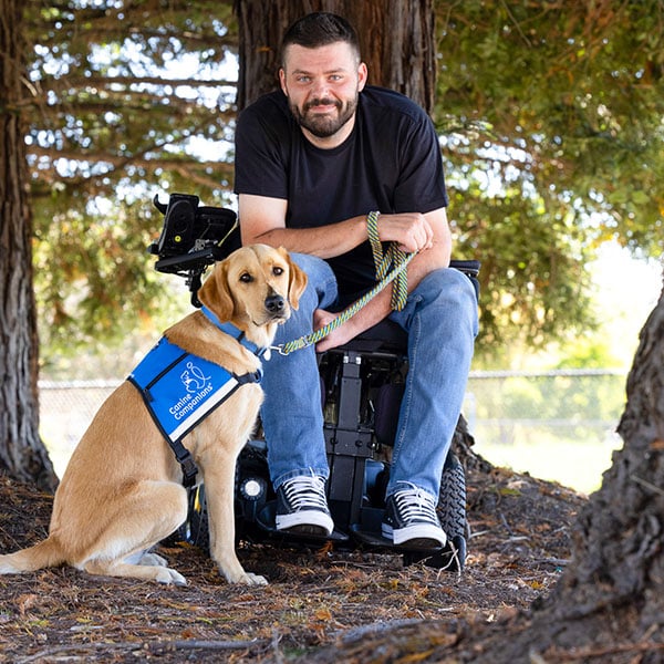 a man in a motorized wheelchair sits next to his yellow lab service dog in a blue service vest in a lush forest