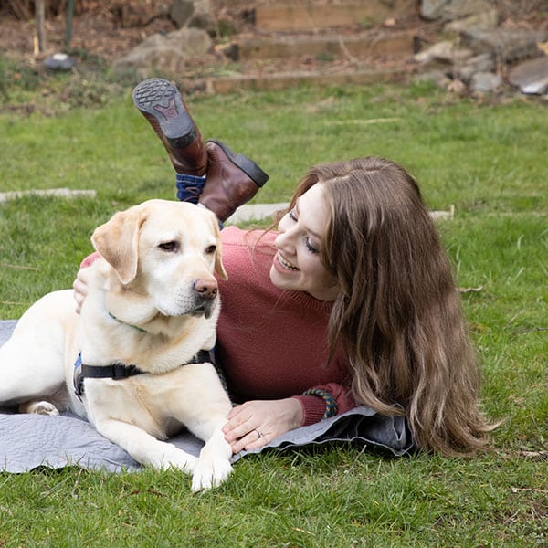 A young woman laying in the grass next to her yellow lab service dog