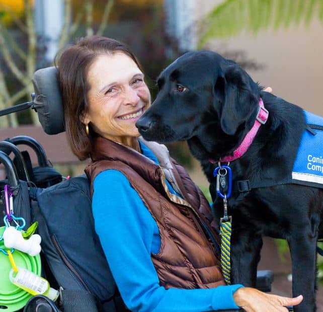 a smiling woman in a motorized wheelchair with a black lab in a blue service vest