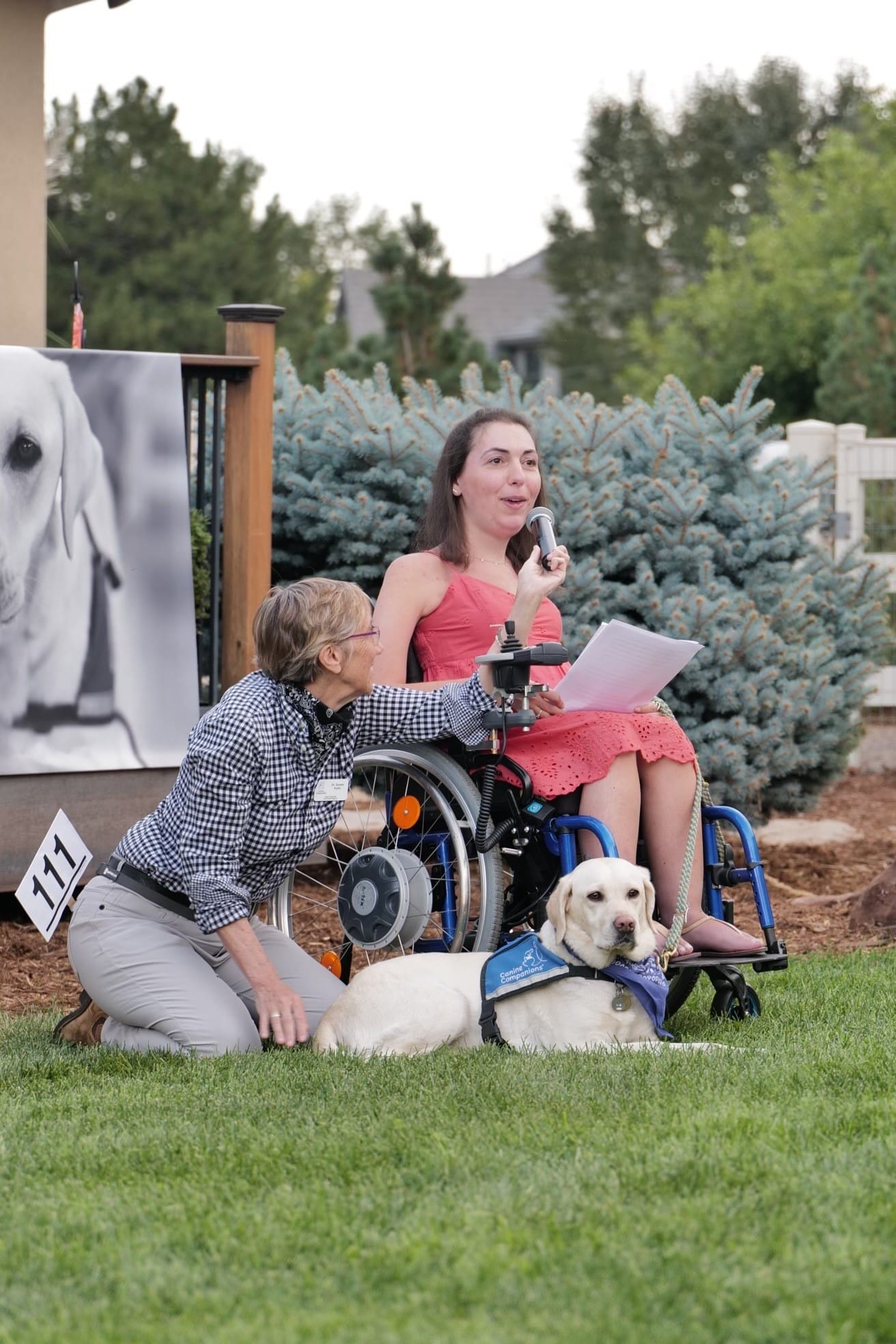 a woman in a wheelchair with a service dog in a blue vest speaks into a microphone