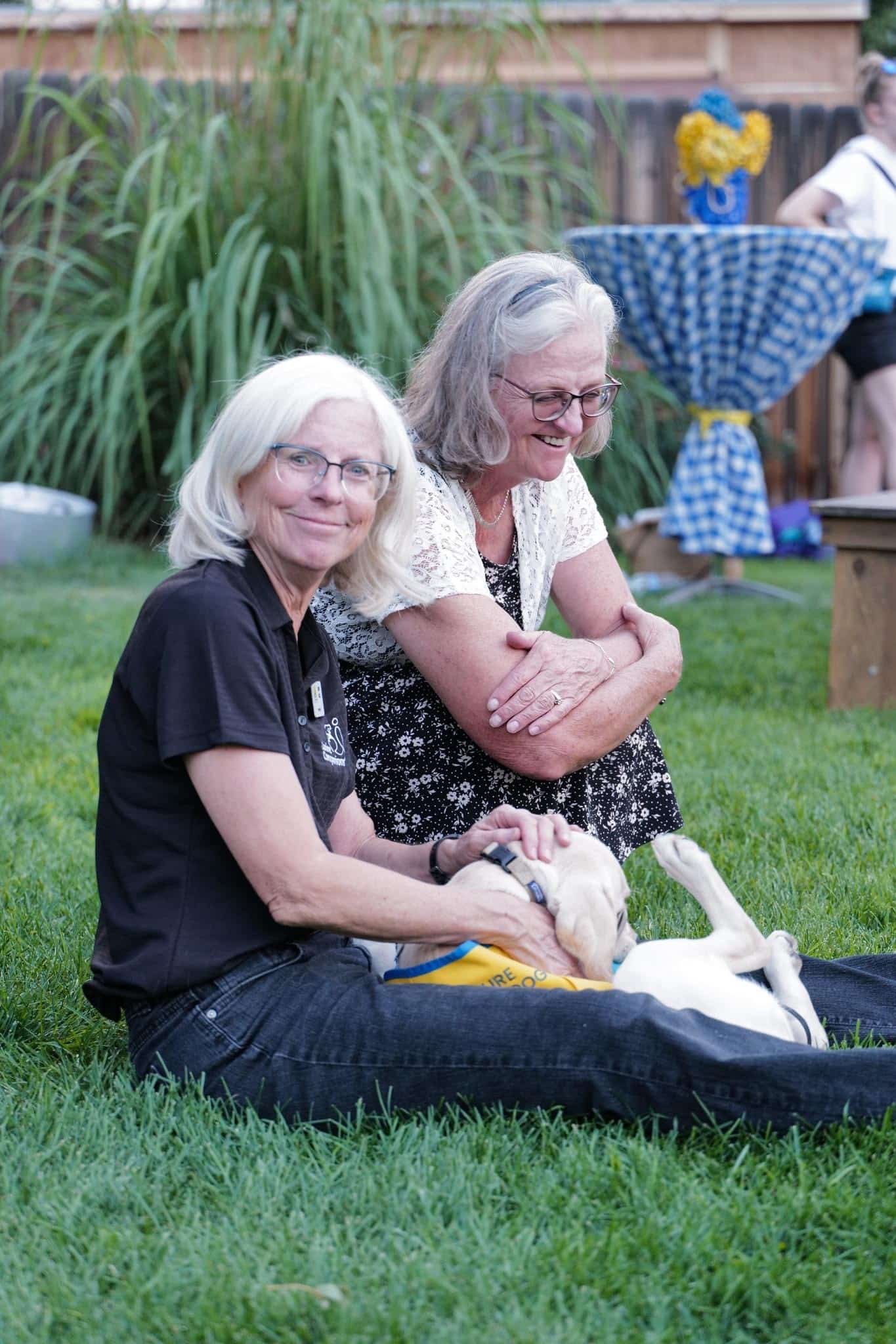 two women sit on a lawn with a yellow lab puppy in a yellow vest