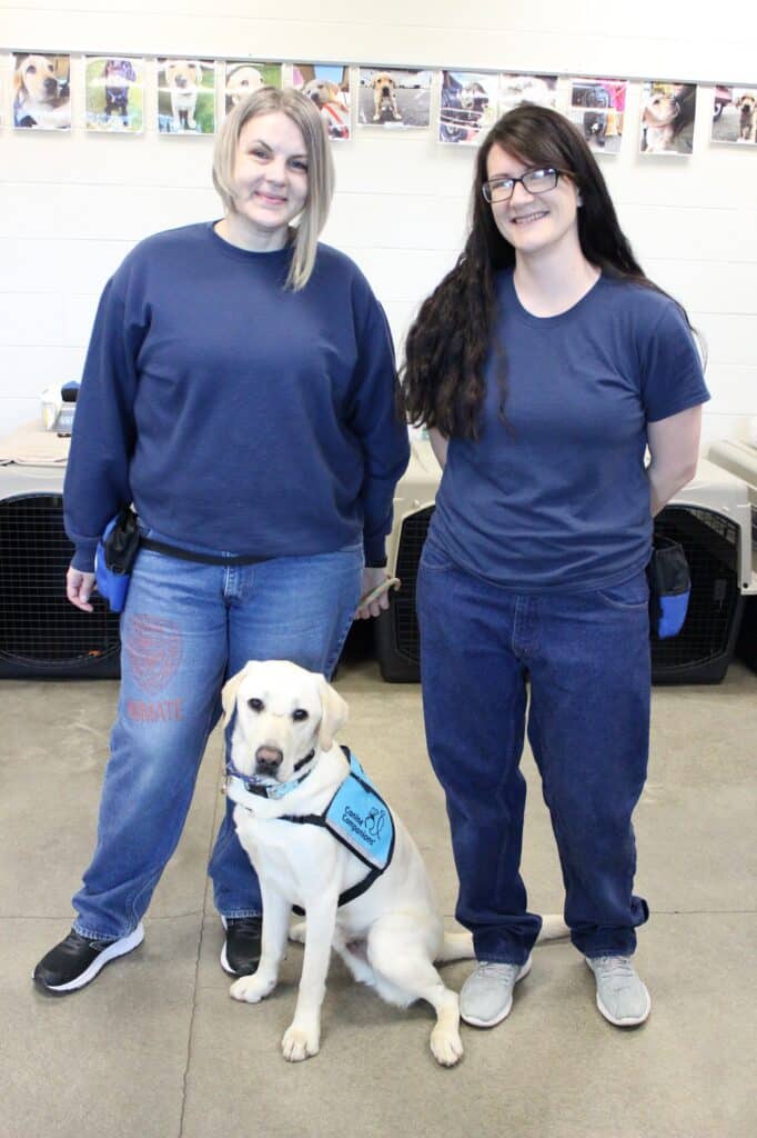 two women stand with a yellow lab in a teal therapy dog vest