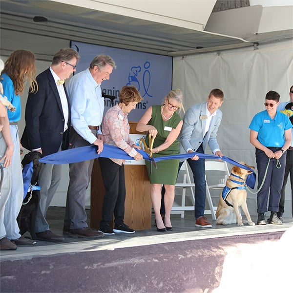 a woman holding a large pair of scissors, cutting a ribbon that’s held by five people standing in a row and two Labradors sitting on each side