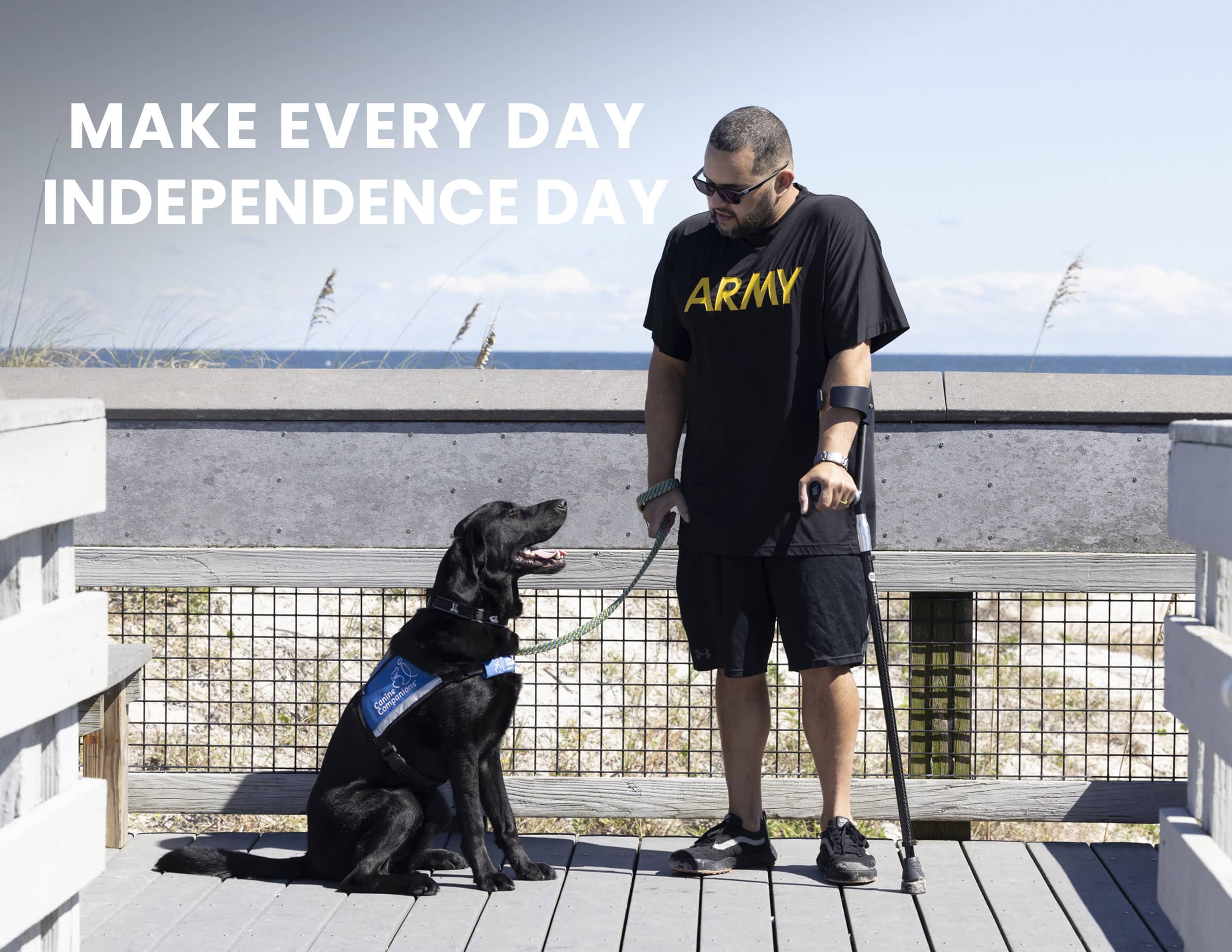 A man wearing an 'ARMY' T-shirt stands with his black service dog on a boardwalk by the beach. The text above them reads 'MAKE EVERY DAY INDEPENDENCE DAY' with the Canine Companions logo at the bottom.
