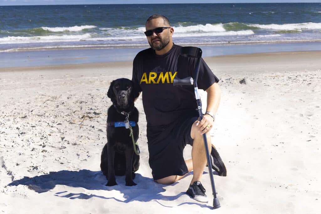 A man wearing an 'ARMY' T-shirt and sunglasses kneels on a sandy beach beside his black service dog, who is sitting next to him, with the ocean waves in the background.