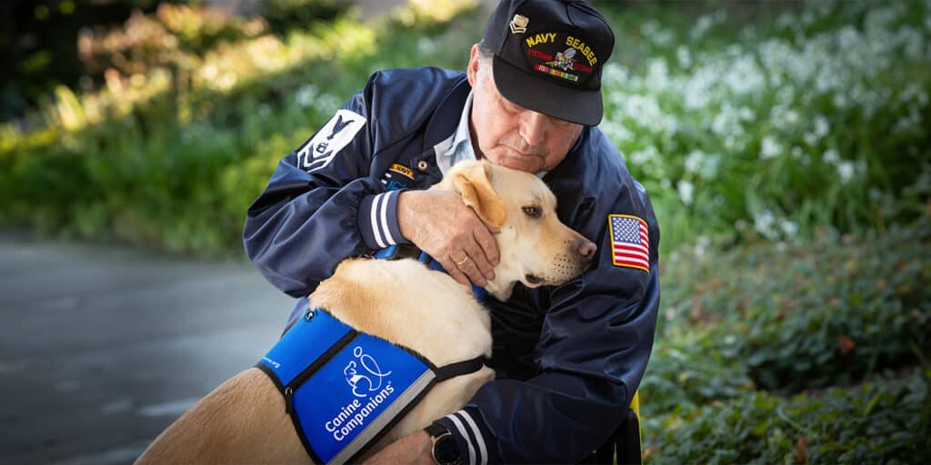 An older man wearing a 'Navy Seabee' hat and a jacket with an American flag patch embraces a yellow Labrador Retriever wearing a blue Canine Companions vest.