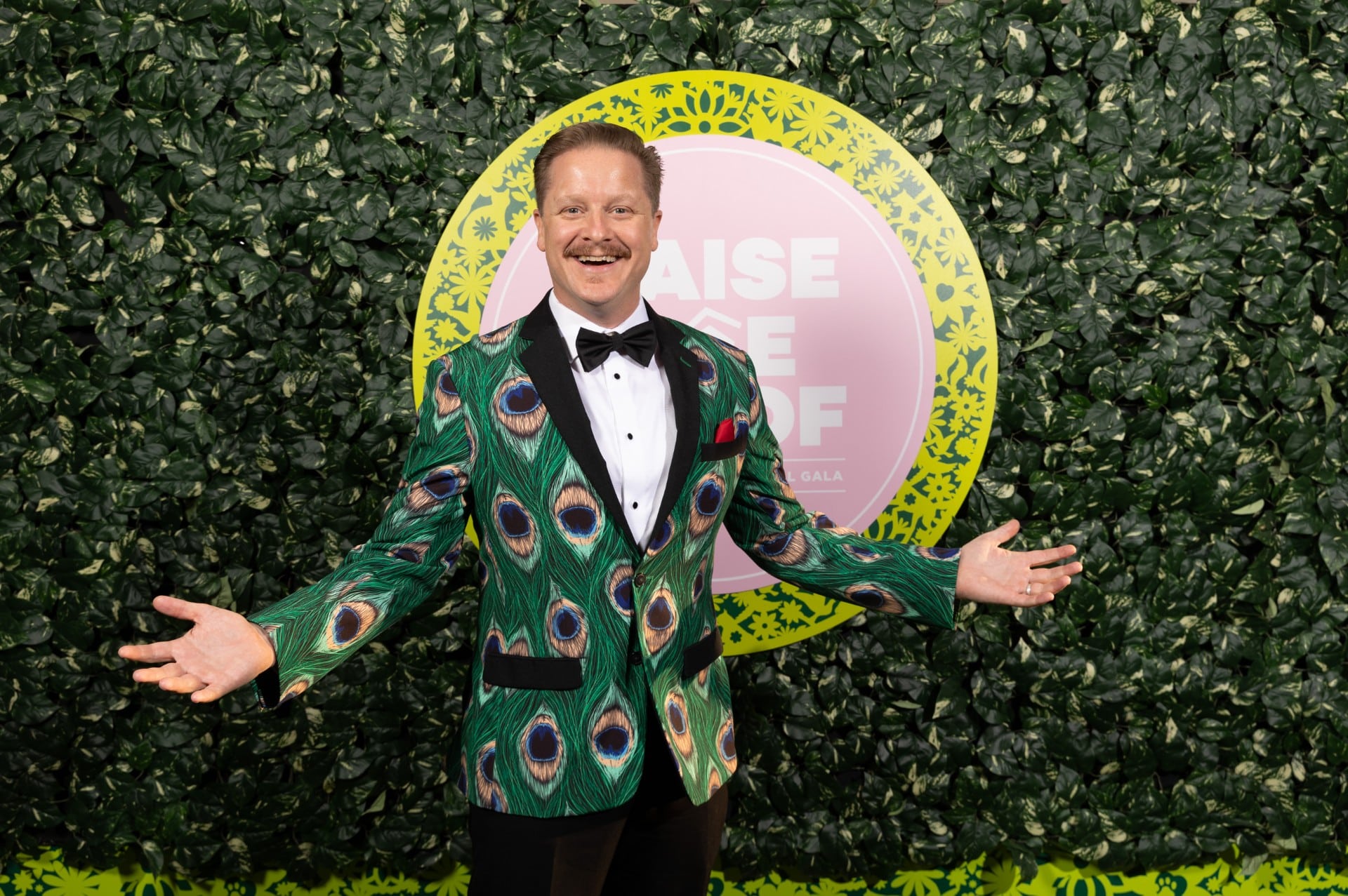 man wearing a peacock jacket standing in front of a leaf wall