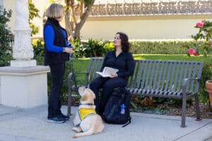 Woman with a yellow puppy wearing a yellow vest talking to a student sitting on a bench