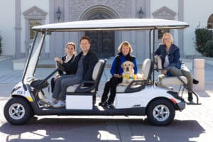 A group of people ride on a golf cart with a puppy in a yellow vest.