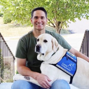 A smiling man with his arm around a yellow lab service dog in a blue canine companions vest