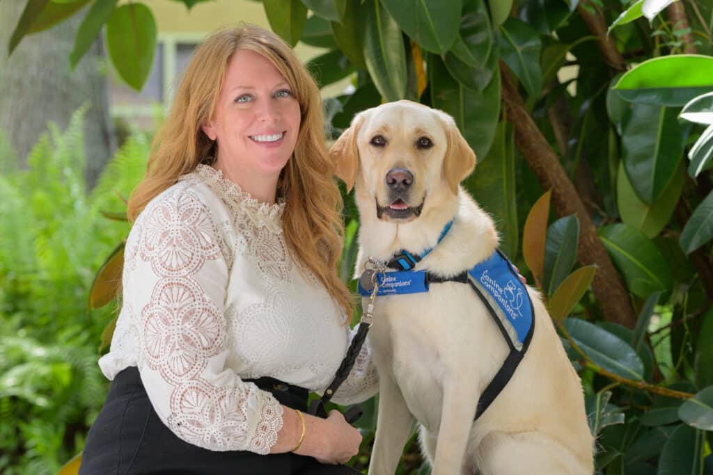 Cathy smiling next to a Labrador in a blue "Canine Companions" vest, outdoors with green foliage.