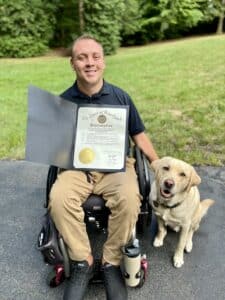 A man in a wheelchair holding an official document, accompanied by a yellow Labrador Retriever.